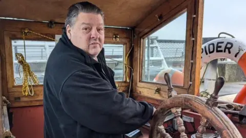 Mal Nicholson Mal is in the steering cabin holding the wheel, and is smiling at the camera. There is a life ring in front of the window that says "Spider"