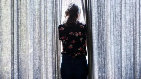 Roos Koole/Getty Images A woman, facing away from the camera, looks out through a pair of grey curtains
