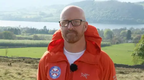 Matt Cox looks at the camera. He is bald with a ginger beard and glasses. Behind him is a large lake and trees. he is wearing an orange jacket with the Patterdale Mountain Rescue team crest on the breast