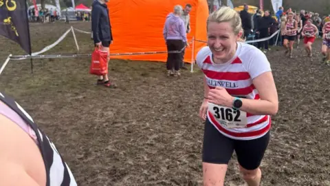 Saltwell Harriers Claire Lloyd smiling while running up a hill. She has blonde hair which is tied up and is wearing a red-and-white striped Saltwell Harriers t-shirt and black shorts. She is also wearing a number bib with 3162 written on it. The tops of her legs are splattered with mud and the ground is completely covered in it. Women are in the background also running up the hill and some spectators stand to the left behind a bit of tape. 