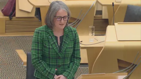 Scottish Parliament Maree Todd wears a green tartan jacket, black top, silver necklace and black-framed glasses with a retainer chain. She is standing in the chamber in Holyrood, with wooden desks and black seats behind her.