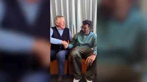 Martin Clunes and Romesh Ranganathan shake hands whilst sat down on an orange sofa.