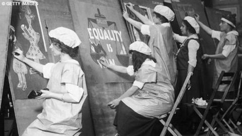 Young girls wearing aprons and caps make equality posters