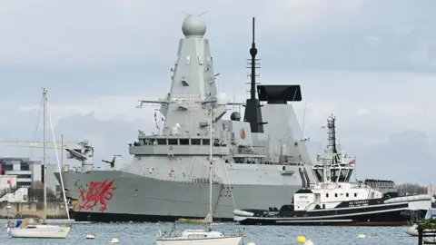 HMS Dragon seen departing Portsmouth Harbour with rocks in the foreground and building behind. We can see the front half of the ship. The red dragon logo on the ship is clearly seen 