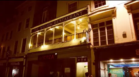 Willow Community Digital Archive/Ceri Oakes Photography An exterior shot of a building lit up at night, with a balcony-style walkway and a large black sign that reads 'The Willow Restaurant'.
