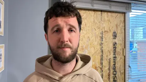 Darian Moore is wearing a beige hoodie. He has short brown hair and and beard. He is standing near the boarded up window in his living room. 