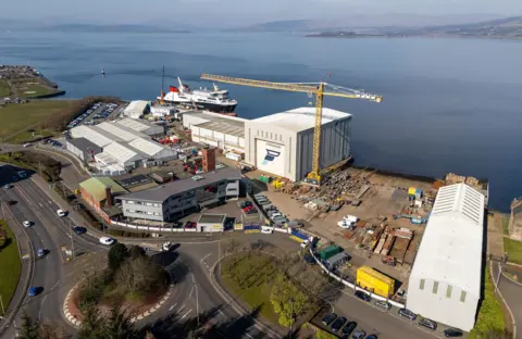 A collection of grey industrial buildings with a yellow crane and a large ferry docked at the quayside