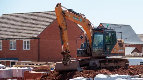 A black and orange digger sits on a large pile of earth surrounded by construction materials with new houses in the background