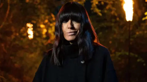 Claudia Winkleman dressed in a black cloak outside with fire on large lamps burning behind her. 