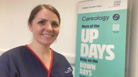 A woman wearing a blue nurse's tunic with red piping standing next to a retractable NHS poster promoting the new app.