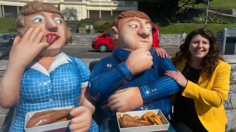 Sarah Gibson has long brown curly hair and is wearing a yellow jacket. She is sitting on a bench next to a sculpture of two people eating boxes of fish and chips