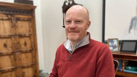 Deputy Gavin St Pier - A man with a bald head, ginger and grey beard, wearing a red jumper and a blue and white shirt.