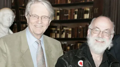 Trinity College Dublin Colin Smythe is wearing a light coloured suit and stood next to Sir Terry who has a white beard and black leather jacket