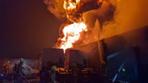 A pillar of smoke and bright flames rise into the night sky from a commercial building. Fire crews are shown in the foreground preparing to tackle the fire.