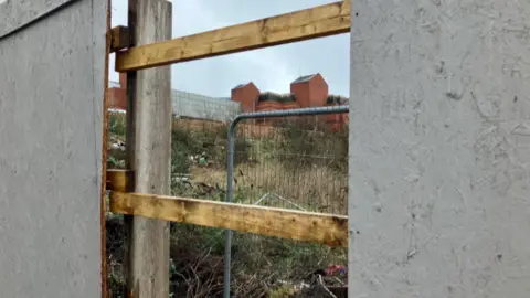 The view through a gap in a grey wooden fence, showing overgrown land.