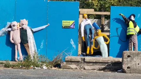 Sara Brady/Copford Scarecrow Festival  Scarecrows resembling Wallace and Grommet, Homer and Marge Simpson and a man in a high vis vest with a speed gun have been nailed to a blue wall 