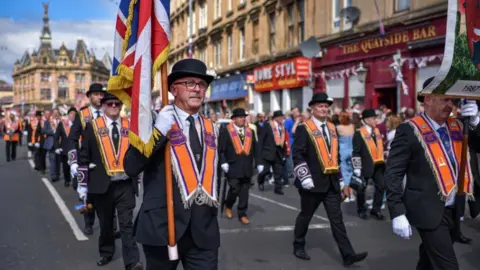 Men in black suits and hats, with orange scarfs draped around their shoulders, march through a street lined by crowds. A man in the foreground is carrying a union flag hanging from a wooden pole. Union flag bunting in the background. 
