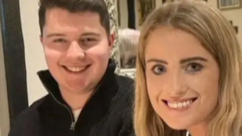 Two people, a man and a woman, sit next to each other smiling at the camera