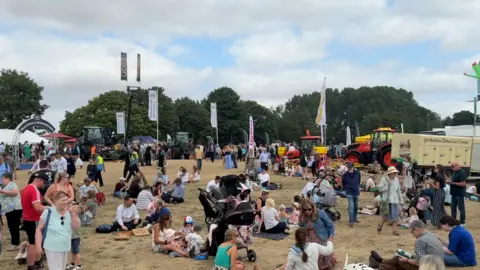Groups of people sat on the grass at Driffield Showground. Tractors can be seen in the background.