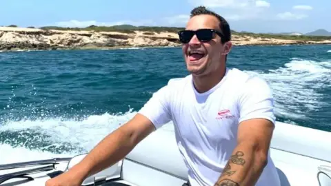 Family photo Ben wearing shades and a white t-shirt whilst steering a speedboat on water on a sunny day