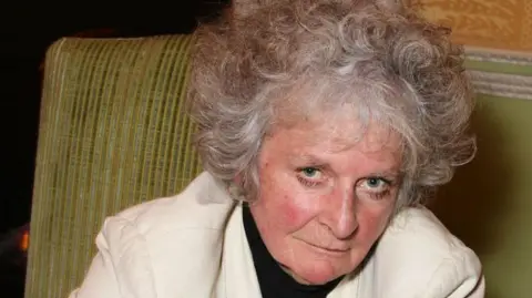 Maggi Hambling looks at the camera and slightly smiles as she leans to the right. She sits on a green sofa. She has short grey curly hair and wears a white jacket with a black top underneath.