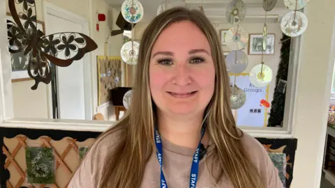 Rachel Britton smiles at the camera in the nursery. She has long dark blonde hair and wears a blue lanyard with 