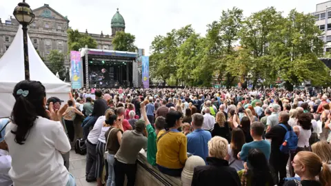 PA Media A large crowd of spectators faces a makeshift stage in the grounds of Belfast City Hall on a bright day. There are trees and tall buildings in the background. 