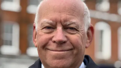 A smiling Jeff Brooks stares straight into the camera. He's wearing a smart blue overcoat over a blue and white striped shirt with a tie in similar colours. Behind him you can just pick out blurred images of closed windows on the upper floors of some of the shops and cafes on Newbury's Market Place.