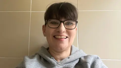 A woman with dark reading glasses and dark hair smiles into the camera. She is wearing a grey hooded top, and has a nose ring.