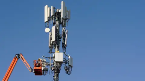 A phone mast in front of a cloudless sky, with an orange maintained crane working on it.