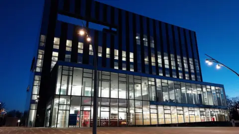 Nadia Lincoln The Corby Cube. A large square building with another smaller one attached in front of it. It has many windows, which are lit up by lights inside the building. The image has been taken at night and so the sky is a very dark blue colour.
