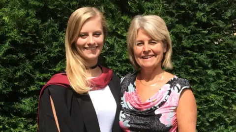 Lucy stands on the left of the picture. She wears a graduation cap and gown. The hood of the gown is burgundy in colour. Joy is standing on the right, in a black cowl-necked dress which is decorated in large pink flowers. Both women are smiling. It is a sunny day, and the pair stand outside beside garden vegetation.