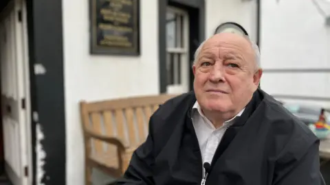 Federica Bedendo/BBC A head-and-shoulder image of George Kemp sitting under a canopy at his pub in Maryport, the Lifeboat. He has short balding white hair and blue eyes. He is wearing a white shirt with a black jacket over him. He is looking at something in the distance with a wistful expression.