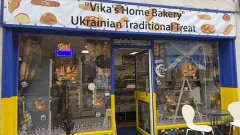 The outside of a bakery. A sign across the top says "Vika's Home Bakery, Ukrainian Traditional Treat".