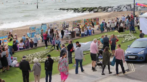 Art on the Prom People walk up and down a coastal promenade where art has been placed on a metal fence for people to enjoy.