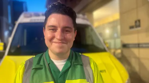 A man with black hair is smiling at the camera. He is wearing a yellow and green hi-vis jacket with silver reflective strips. He is standing in front of a yellow and white ambulance vehicle