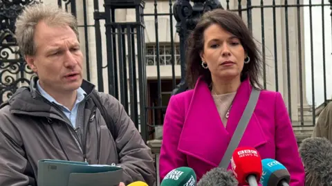 Daniel Holder, Gráinne Teggart and Mark Thompson outside Laganside court in Belfast. There is a collection of branded microphones in front of them. Daniel is holding folders and wearing a grey coat. Grainne is wearing a pink coat and has a bag over her shoulder. Mark is holding a poster and wearing a green coat.