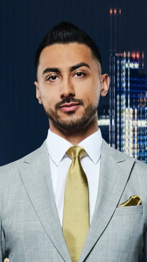A young man with a beard wearing grey suit and gold tie