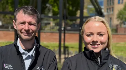 A woman with blonde long hair and a man with short spicky brown hair smiling at camera. They are both wearing black jackets with logos of Causeway Coast and Glens Borough Council and 3GS. Sarah's jacket has a camera attached. There is a park in the background with people standing talking blurred in the background.
