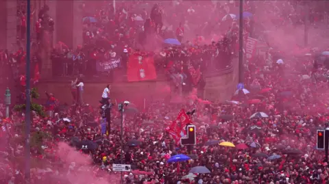 PA A large group of Liverpool fans crowd the street with some people sat on traffic lights, others have climbed onto buildings