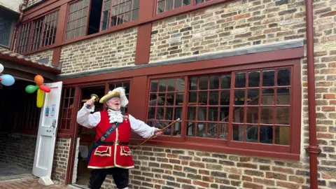 A traditional English town crier in a red and gold tabbard stands outside an old brick building with dark red sash windows. He is holding a gold bell, wearing a feathered three-corner hat and is mid-cry.