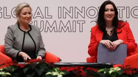 PA Media O'Neill (left) is sitting on a red chair, wearing a grey dress and matching blazer. Her blonde hair is tied up, Little-Pengelly is sitting on a red chair and is wearing a black dress and red blazer. She has long dark brown hair.