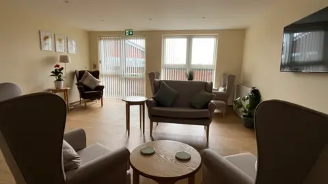 A room with six xhairs on a wooden floor with windows with blinds on the far wall and flowers on a low table