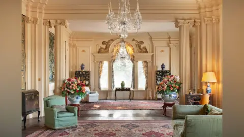 Trent Park Museum Trust A large state room with furniture, expensive rugs and fine objects. There is a large crystal chandelier. 