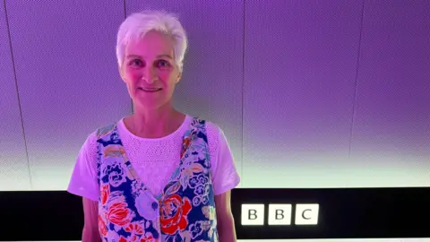 Fiona is standing in front of a purple background in a BBC building. She has short white hair and is wearing a white T-shirt under a blue, white and red tunic dress.