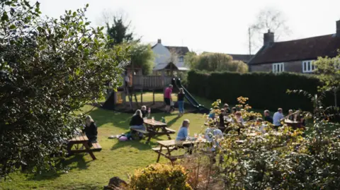 The Waldegrave An out of focus garden with a children's play castle and picnic benches. In the foreground are bushes in focus.