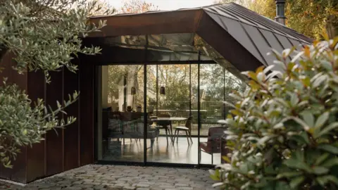 RIBA The image shows one of the rooms behind a glass front. A dining table, chairs and lamps can be seen through the floor-length windows.