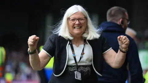 Port Vale Football Club owner Carol Shanahan is seen wearing glasses, a lanyard, white blouse and black jacket with white piping. She is clenching her fists and smiling in celebration.