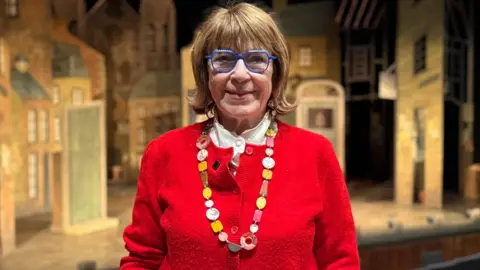 A woman standing in the Lyric Theatre. She is wearing a red jumper, colourful necklace and white shirt. She has brown hair and purple glasses. The stage is behind her.