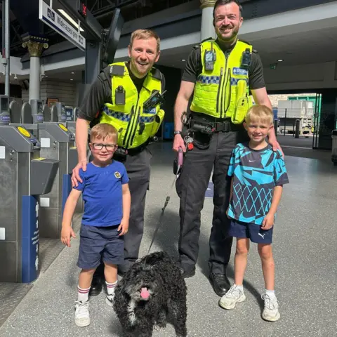 Lauren Stirling Two male police officers standing with two young boys holding a black dog on a lead.